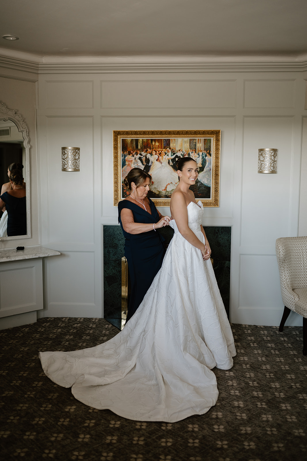Mother of the bride helping fasten the wedding gown as the smiling bride looks back over her shoulder.